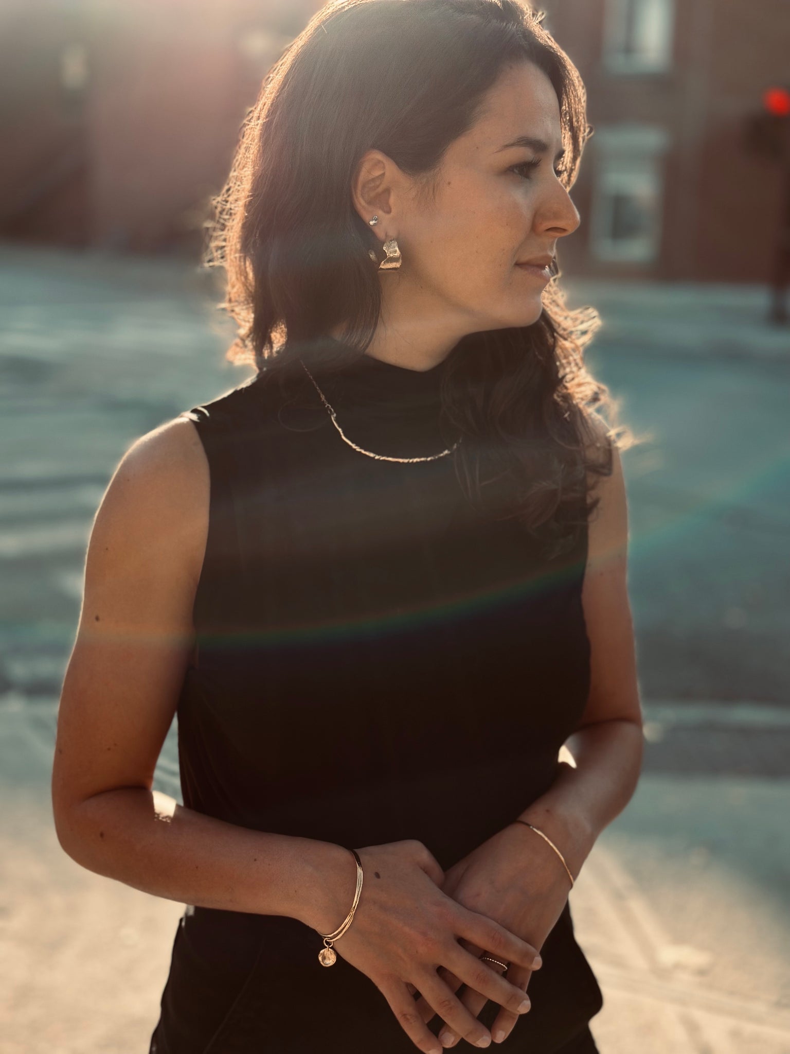 Woman in sunset light wearing a black sleeveless top, gold charm earrings, and a delicate gold bracelet.