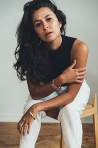 Woman in chic black sleeveless top and white pants with gold jewelry, posing thoughtfully on wooden chair.