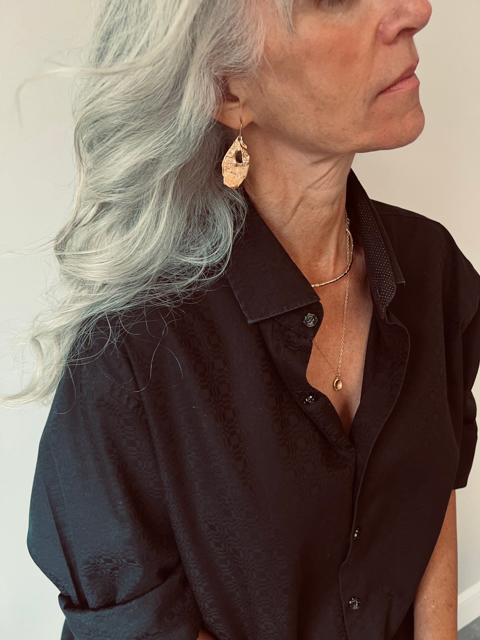 Side profile of a woman with silver hair wearing a textured gold leaf earring and a layered gold necklace with a black shirt.