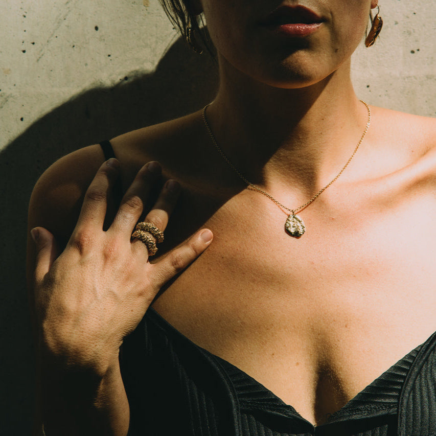 Woman wearing a black top with a gold necklace and gold ring,made by what if you stayed, standing against a concrete wall.
