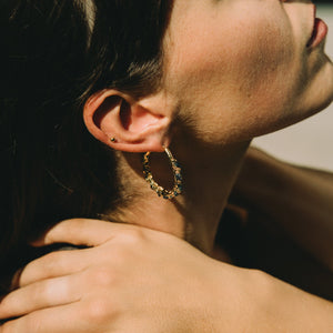 Close-up of a person with their head resting on another person's shoulder, with a soft focus background, wearing large gold hoop earrings with blue raw sapphires made by what if you stayed
