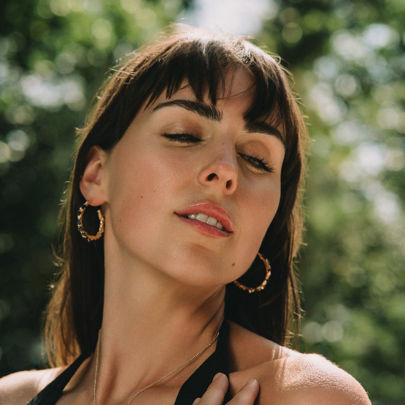 Woman with gold hoop earrings and a necklace outdoors with greenery in the background