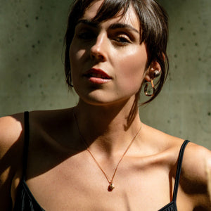 Woman against a textured wall wearing a chain pendant necklace in gold with a drop shape pendant, textured organic rings in gold with precious stones and gold earrings made by what if you stayed 