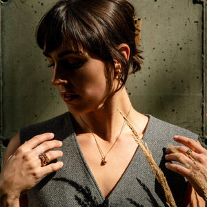 Woman holding a wheat stalk against a textured wall wearing a chain pendant necklace in gold with a drop shape pendant, textured organic rings in gold with precious stones and gold earrings made by what if you stayed 