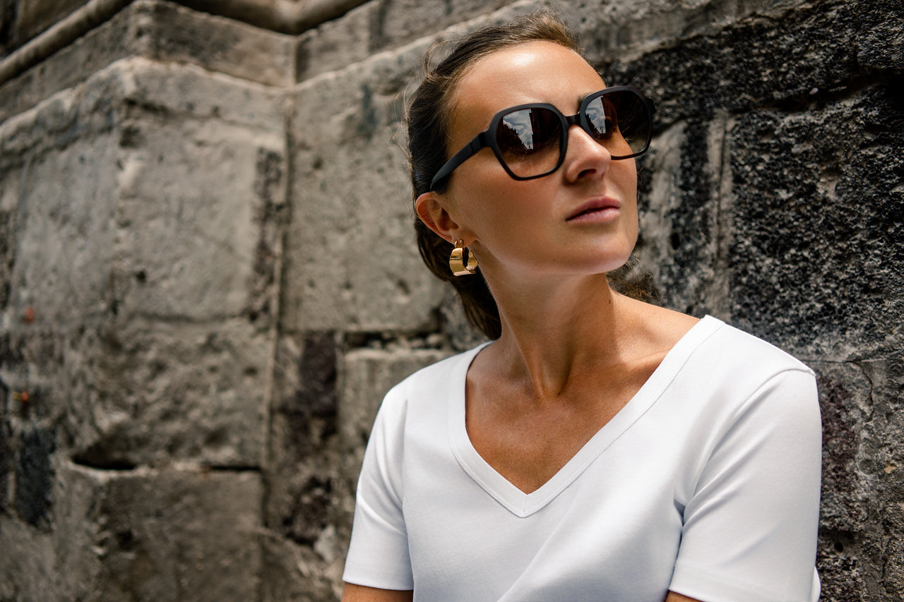 Woman wearing sunglasses and a white top against a stone wall wearing gold large eclipses earrings made by what if you stayed
