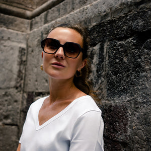 Woman wearing sunglasses and a white top against a stone wall wearing gold earrings hoops made by what if you stayed 