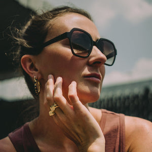 Woman wearing sunglasses and gold earrings with raw sapphires and large gold textured rings made by what if you stayed with a blurred background