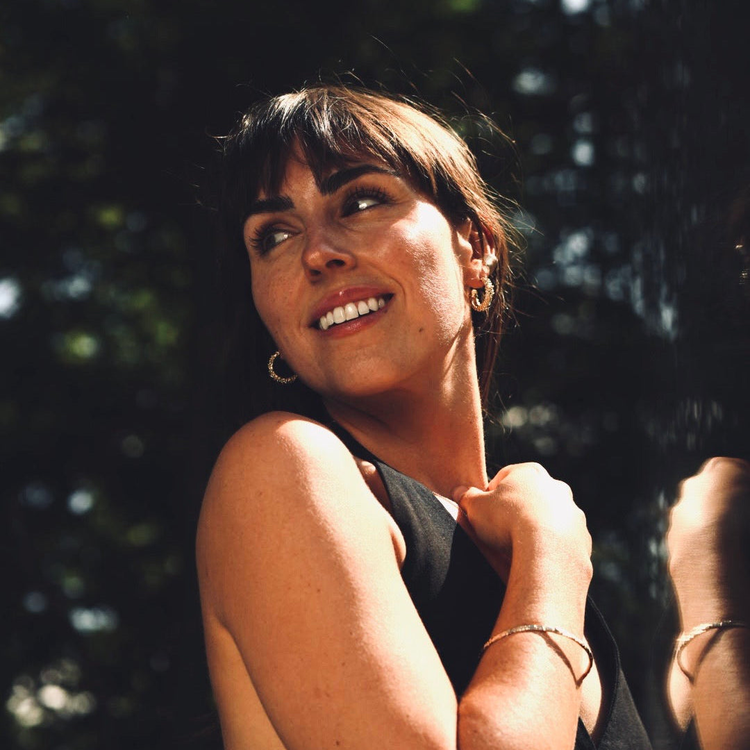 Woman with dark hair smiling outdoors with a blurred background wearing gold jewelry made by what if you stayed
