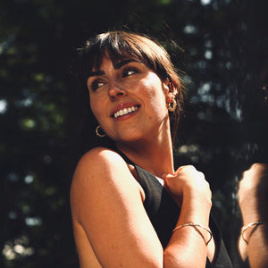 Woman with dark hair smiling outdoors with a blurred background wearing gold jewelry made by what if you stayed