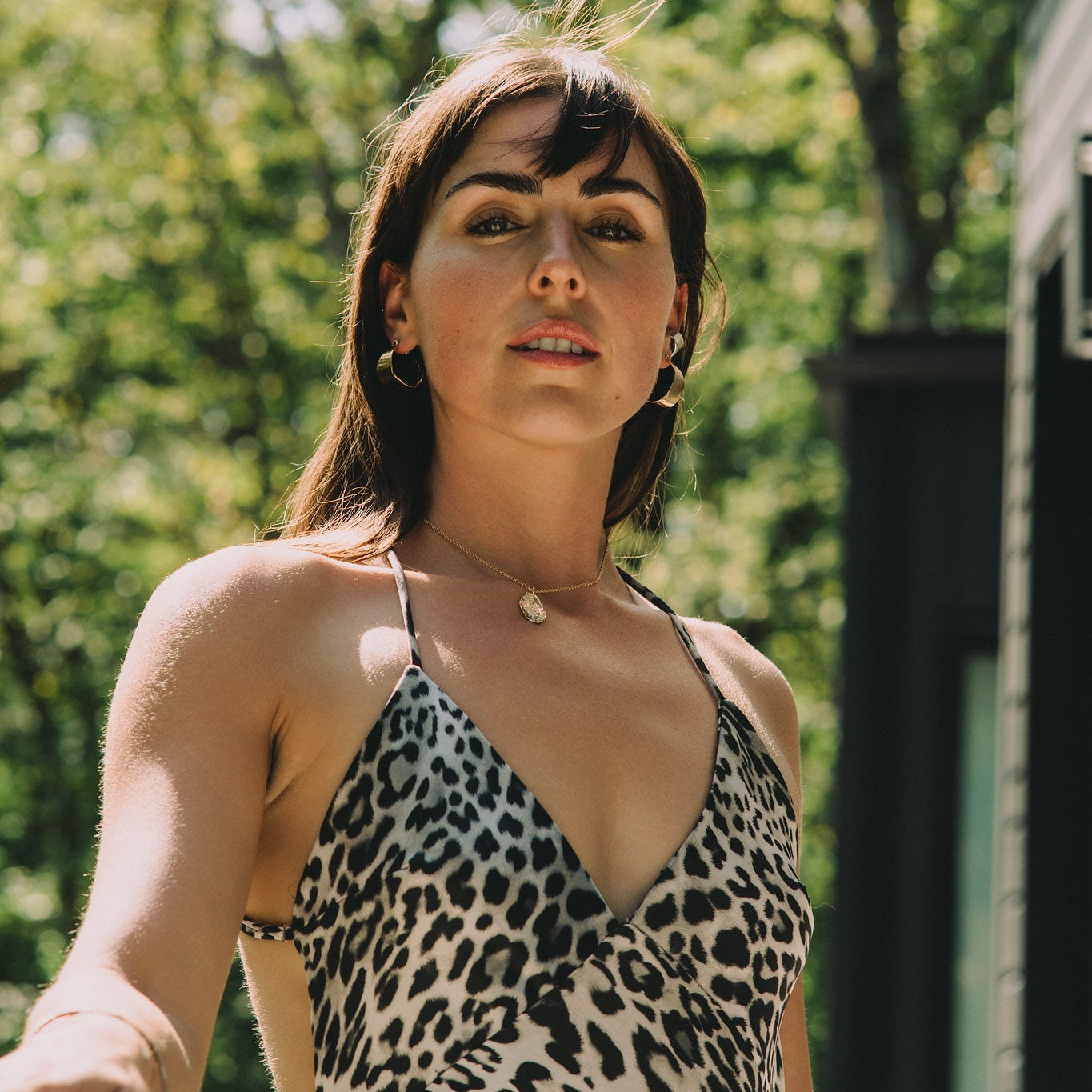 Woman wearing a leopard print dress outdoors with greenery in the background wearing large gold earrings and gold pendant made by what if you stayed 