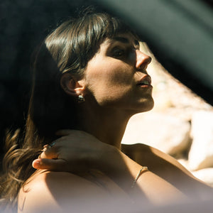 Woman looking out of a car window with sunlight filtering through, wearing what if you stayed bangle in gold and minimalist earrings hoops in gold