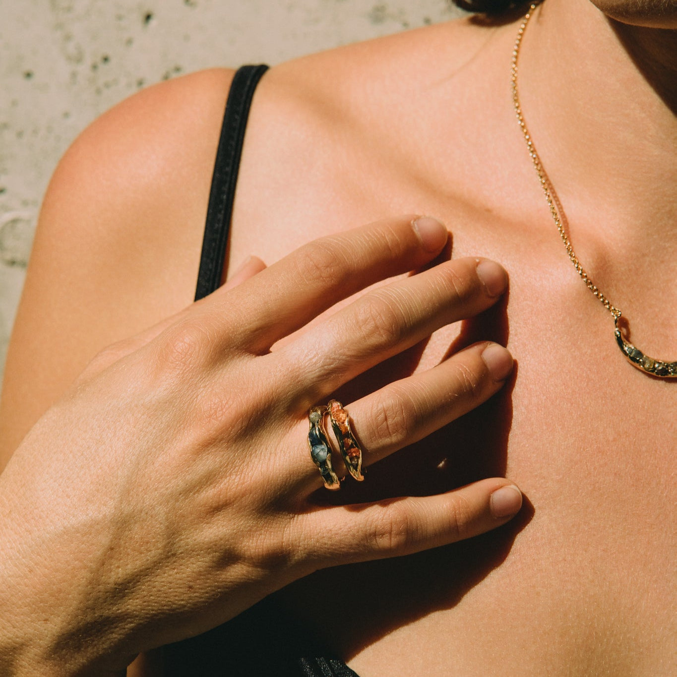 Young woman wearing a black top against a concrete wall in the sun light, wearing gold rings with raw stones and a gold necklace made by What If You Stayed